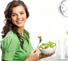 mujer joven sonriendo mientras sostiene en una mano un plato con ensalada y en la otra un tenedor