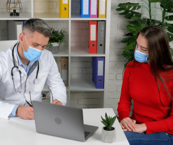 médico con mascarilla y detalantal blanco escribe en su escritorio mientras conversa con paciente mujer joven, de cabello largo y polera roja