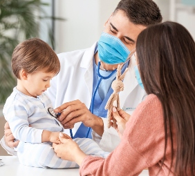 médico con mascarilla examina a niño pequeño que está sobre una mesa mientras su mamá le toma la mano